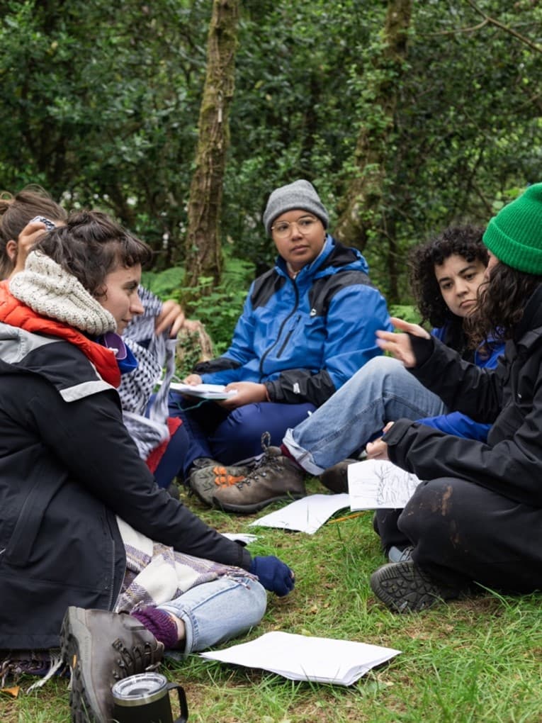 People sat in winter clothes on grass talking to each other with paper in front of them.