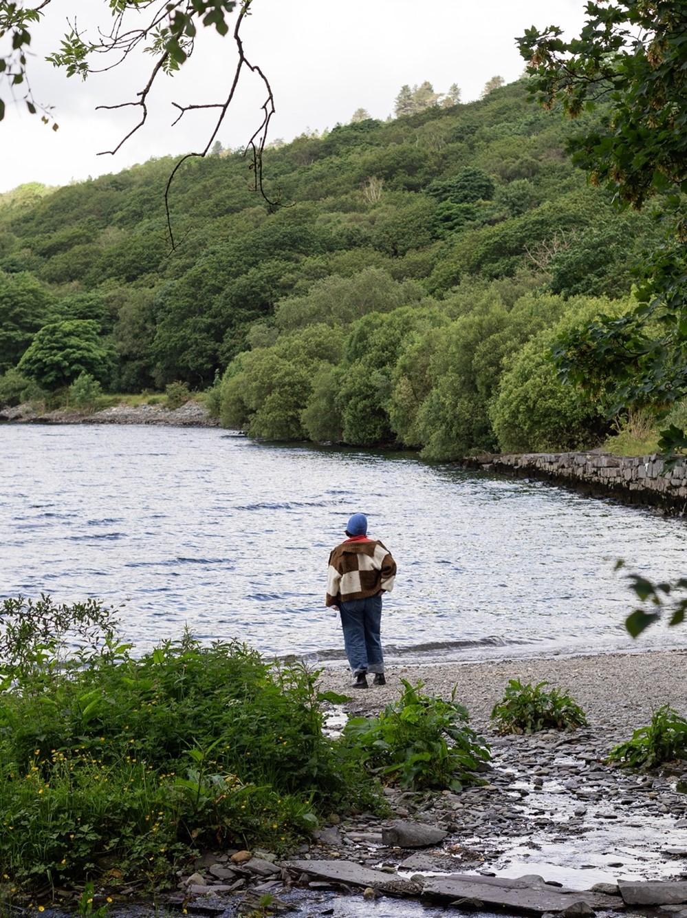 Somebody standing on a small rocky beach by a lake surrounded by woodland.