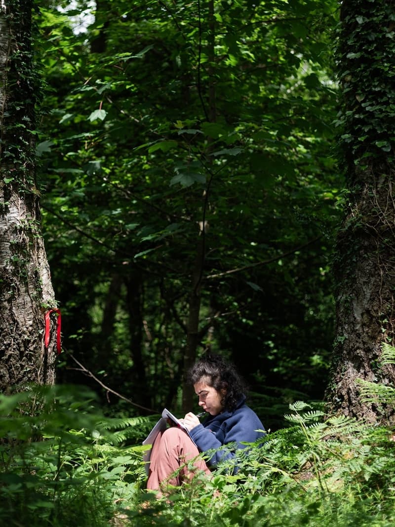 A person sat under a tree resting paper on their knees and writing.