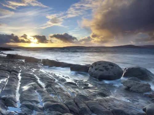 A coastal landscape, with the sea gently lapping at smooth rocks as the sun sets behind scattered clouds