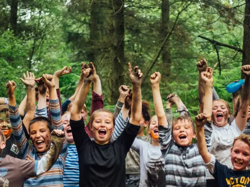 Children with their hands in the air and paint on their faces in a woodland